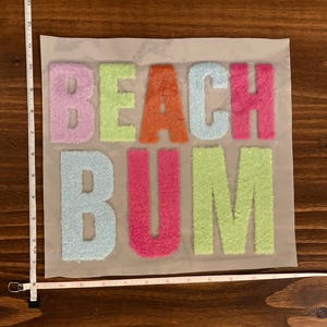 May include: A colorful, textured patch with the words "BEACH BUM" in vibrant letters. The letters are in shades of pink, orange, green, and blue, creating a playful and eye-catching design. The patch is likely for clothing or accessories.