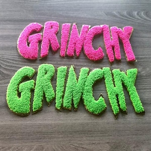 May include: Two words, "GRINCHY", are displayed in the image. One word is in bright pink, the other in green. Both words have a textured, fuzzy appearance with a gold border. The words are placed on a gray wooden surface.