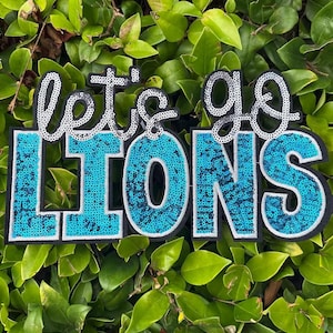May include: A sequined applique with the words "let's go LIONS" in silver and turquoise. The letters "LIONS" are in a bold, block font, filled with turquoise sequins. The words are outlined in black and white, set against a backdrop of green leaves.