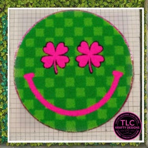 May include: A round, green doormat with a checkered pattern and a smiling face design. The eyes are two pink shamrocks, and the mouth is a curved pink line. The doormat has the text "TLC Krafty Designs" in a circular logo.
