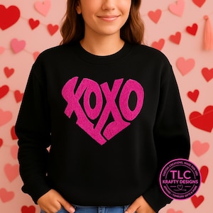 May include: Black sweatshirt with a large pink heart design. The heart is filled with the letters "XOXO". The sweatshirt has a crew neck and long sleeves. The design is centered on the front of the shirt. The brand logo is on the lower right side.
