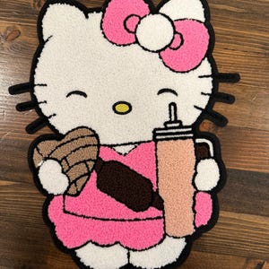 May include: A white, pink, and black rug featuring a cartoon cat character holding a drink and a pastry. The cat has a pink bow, a pink dress, and a brown bag. The rug has a black border.