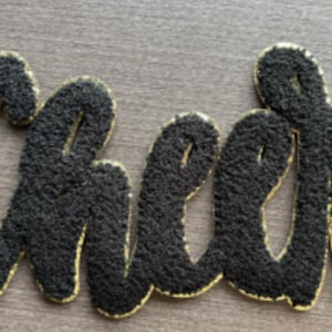 May include: A black, textured "Cheer" word art sign with gold trim. The letters are in a cursive font and have a fuzzy, plush appearance. The sign is likely used for home decor or celebrations.