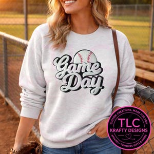 May include: A light gray sweatshirt with the words "Game Day" in a script font, with a baseball graphic. The sweatshirt has long sleeves and a crew neck. The image also includes a logo that says "TLC Krafty Designs".