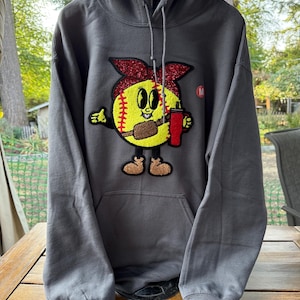 May include: A charcoal gray hoodie with a front pocket and a drawstring hood. The hoodie features a colorful patch of a baseball character holding a red cup. The character has a yellow face, brown shoes, and a red bow.