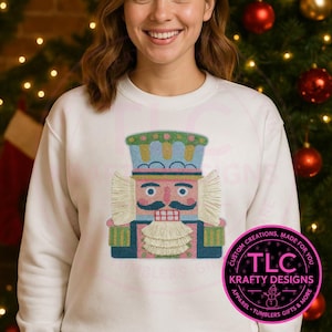May include: White sweatshirt featuring a colorful embroidered nutcracker design. The nutcracker has a pink face, blue hat, and a white beard. The design includes the text "TLC Krafty Designs" in a circular logo.