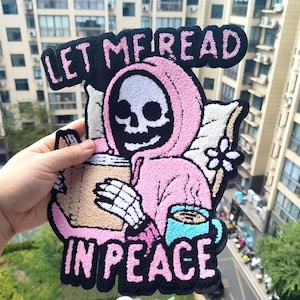 May include: Embroidered patch featuring a skeleton in a pink hooded robe, holding a coffee cup and a blue mug. The patch has the text "LET ME READ IN PEACE" in pink and white. The design is set against a black background.