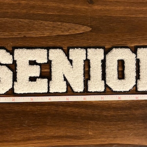 May include: A white and black chenille patch that spells out the word "SENIOR". The patch is approximately 10 inches long and is placed on a wooden surface. The letters are white with a black border.