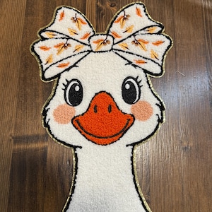 May include: A white goose-shaped rug with a large bow featuring orange and yellow leaf accents. The goose has a bright orange beak, rosy cheeks, and black eyes. The rug is outlined in gold and has a textured, plush appearance.