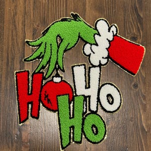 May include: A Christmas-themed applique featuring a green hand holding a red ornament and a red sleeve with white trim. The words "HO HO HO" are in red, white, and green, outlined in black and gold. The design is on a wooden surface.