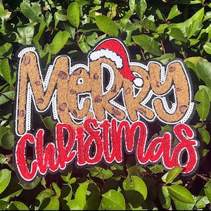 May include: A festive sign with the words "MERRY CHRISTMAS" in a decorative font. The word "MERRY" is in a brown, glittery font with a Santa hat on top. "CHRISTMAS" is in red with a white outline.