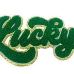 May include: A green felt patch with the word "Lucky" in a script font. The letters are a dark green color with a gold glitter outline. The patch is likely for clothing or accessories.