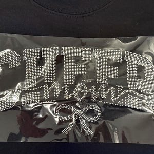 May include: Black t-shirt with a rhinestone design that reads "CHFFD Mom" with a rhinestone bow. The design is on a clear plastic sheet, ready for application. The shirt is made of a soft, dark fabric.