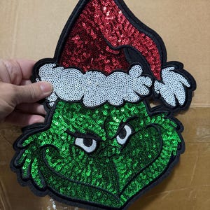 May include: A sequined Grinch face patch with a red Santa hat and white trim. The Grinch's face is green with white eyes and a black outline. The patch is held by a hand.