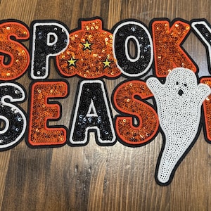May include: A Halloween-themed patch with the words "Spooky Season" in black and orange sequined letters. A white sequined ghost is on the right. The pumpkin-shaped "O" has yellow star accents.