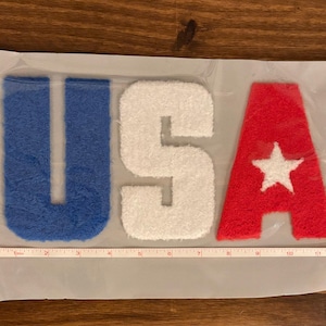 May include: A patriotic patch featuring the letters "USA" in blue, white, and red. The "U" and "A" are approximately 4 inches tall, and the "S" is about 4 inches tall. The red "A" has a white star. The patch is on a mesh backing.