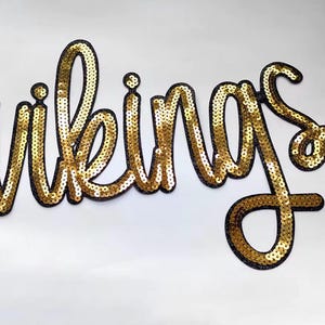 May include: Gold sequined "Vikings" script. The word is outlined in black and sits on a white background. The sequins are densely packed, creating a shimmering effect. The letters are in a cursive style.
