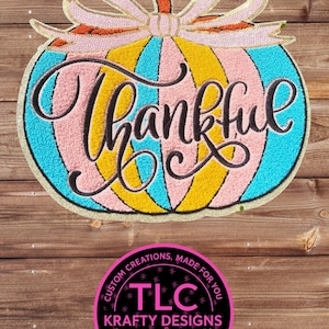 May include: A pumpkin-shaped patch featuring alternating vertical stripes of turquoise, yellow, and pink, topped with a pink sequined bow. The word Thankful is embroidered in black script across the center. Below is a circular black logo with pink text reading TLC KRAFTY DESIGNS CUSTOM CREATIONS MADE FOR YOU APPAREL TUMBLERS GIFTS & MORE.