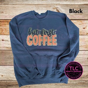 May include: A navy blue sweatshirt with the text "But first COFFEE" in a glittery orange and green design. The word "Black" is in the upper right corner. A circular logo is in the lower right corner.
