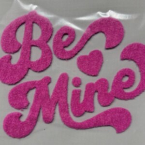 May include: A pink, fuzzy felt applique with the words "Be Mine" in a cursive font. A small heart shape is between the words. The letters are a vibrant pink color, and the applique is likely for crafting or decorating.