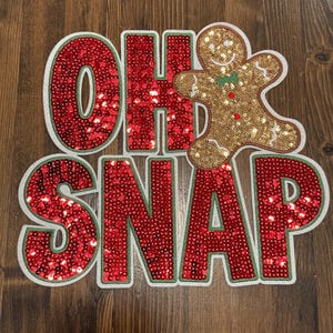 May include: A Christmas decoration with the words "OH SNAP" in red sequins with a green border. A gold sequined gingerbread man with a green bow tie is next to the word "OH".