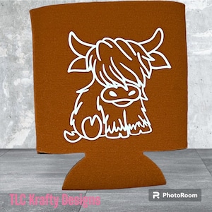 May include: A brown drink koozie featuring a white outline of a Highland cow. The koozie has a rectangular shape with a cutout at the bottom. The text "TLC-Krafty Designs" is visible at the bottom left.