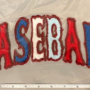 May include: A baseball-themed patch with the word "BASEBALL" in a curved design. The letters are red, white, and blue with a brown border. The patch is on a light gray background.