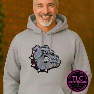 May include: A gray hooded sweatshirt featuring a sparkling bulldog graphic. The bulldog design is in shades of silver, purple, and black. The sweatshirt has a front pocket and a drawstring hood. The text "CUSTOM CREATIONS, MADE FOR YOU" and "TLC KRAFTY DESIGNS" are also visible.