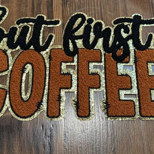 May include: A decorative sign with the phrase "but first COFFEE" in a bold, embroidered style. The text is in black and brown with a gold glitter outline, set against a wood grain background. A coffee-themed item.