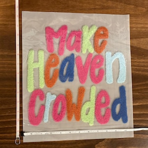 May include: A colorful textile piece with the phrase "Make Heaven Crowded" in vibrant, textured letters. The letters are in shades of pink, orange, blue, green, and light blue. The piece is approximately 10 inches wide.