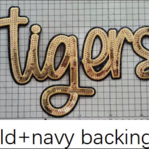 May include: A script-style patch featuring the word tigers in gold sequins with a navy blue border. The patch is positioned on a white grid mat that shows measurements in centimeters, with the text gold+navy backing printed below the image.