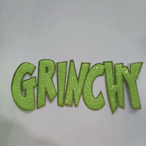 May include: Lime green sequined letters spell out the word "GRINCHY". The letters are outlined in a darker green and are slightly angled. The background is a plain white.