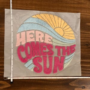 May include: A colorful, circular patch with the text "HERE COMES THE SUN". The design features a yellow sun, blue and green waves, and pink lettering. The patch is approximately 12 inches wide.