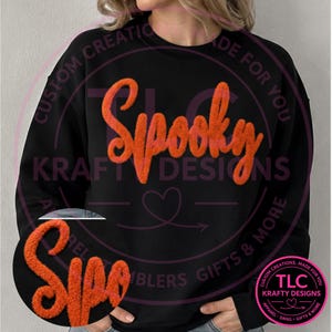 May include: A black long-sleeve sweatshirt featuring the word Spooky in large, textured orange chenille letters across the chest. The fabric has a soft, fuzzy appearance, and the design includes a small heart outline below the text.