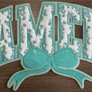 May include: A large iron-on patch featuring the word AMEN in block letters. The letters have a white textured surface with a light blue floral pattern and a light blue border. Below the letters is a large, bright light blue sequined bow.