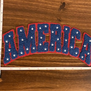 May include: A blue and red felt patch with the word "AMERICA" in white star-filled letters. The patch is approximately 7 inches wide and 3 inches tall. The patch has a red border and is on a wooden surface.