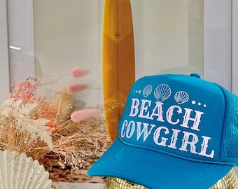 May include: A turquoise blue trucker hat with a white mesh back. The hat has white embroidered text that says "Beach Cowgirl" and three white seashells.