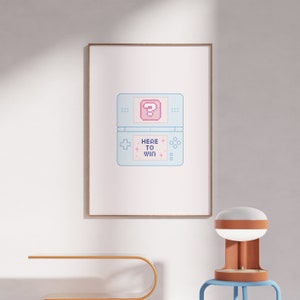 Könnte beinhalten: Ein gerahmter Druck eines pixelartigen Nintendo DS mit einem Fragezeichenblock auf dem Bildschirm und dem Text "HERE TO WIN". Der Druck ist auf weißem Hintergrund und hat einen hellblauen Rahmen.