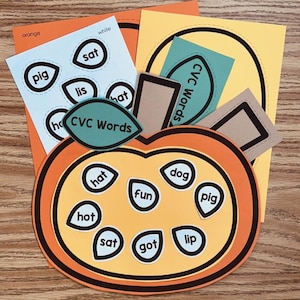 May include: A colorful educational activity featuring a pumpkin-shaped cutout with CVC words. Includes orange, yellow, and blue paper elements with words like "hat," "dog," and "pig." The words are in a white font.