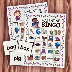 Op de afbeelding: Valentijnsdag Bingo-spel met kleurrijke illustraties van een kat, een varken, een huis, een vis, een doos, een net, een ventilator, een maïskolf, een hart, een kip, een schildpad, een tas, een bed, een bot en een honkbalknuppel. Het spel bevat oproepkaarten met de woorden "tas", "doos" en "varken".
