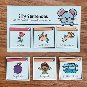 May include: Educational game with cards for creating silly sentences. The cards feature illustrations and text, including a flower, clapping hands, and a plum. The set includes a mouse illustration and the text "Silly Sentences".