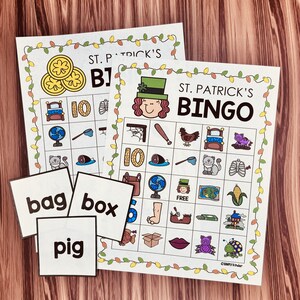 May include: Two printable St. Patrick's Day Bingo cards with images of a leprechaun, a pot of gold, a shamrock, a cat, a fish, a net, a bed, a pig, a corn cob, a house, a fan, a foot, a shoe, a  lipstick, a spider, a ladybug, and a  number 10.  The cards are surrounded by a garland of lights.  There are also three calling cards with the words "bag", "box", and "pig".