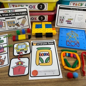 May include: A set of educational activity cards for preschoolers. The cards feature colorful illustrations of a school bus, a house, a snake, a birthday cake, a dog, and a crayon box. The cards are labeled "Task Box" and include instructions for completing the activities. The set also includes a blue plastic frame with colorful rubber bands, a set of colorful play dough, and a pair of red tongs.