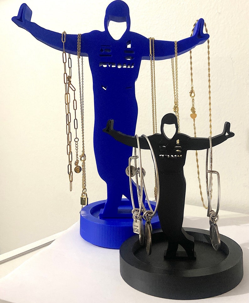 May include: A blue and black jewelry stand shaped like two figures. The larger figure is holding necklaces and the smaller figure is holding bracelets. The stand has a round base.