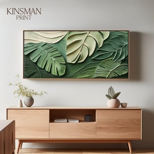 May include: A rectangular framed canvas print with a 3D textured design of tropical leaves in shades of green and cream. The artwork is mounted in a light wood frame. The text "KINSMAN PRINT" is visible in the upper left corner.