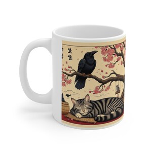 May include: White ceramic mug with a black crow perched on a branch of a cherry blossom tree. The crow is facing right and has a black beak. The branch is covered in pink blossoms. A gray and white striped cat is sleeping on a red mat below the branch. The text "春" is written in black on the mug.