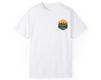 May include: White t-shirt with a small, colorful patch on the upper left chest. The patch features a mountain scene with the words "CONFIDENT AND UNASHAMED" in a stylized font. The shirt is made of a soft, likely cotton material.