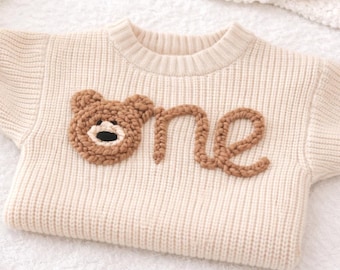 May include: Cream-colored baby sweater with a ribbed knit texture. The sweater features a brown bear applique and the word "one" in brown script. The sleeves have ribbed cuffs. The sweater is laying on a white surface.