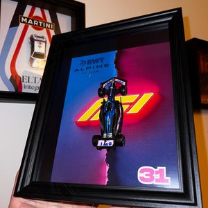 Puede incluir: Obra de arte enmarcada con un modelo de coche de carreras de Fórmula 1 azul y negro. El fondo tiene un degradado azul y rosa con el número 31 en rosa. El marco es negro.