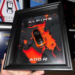 Könnte beinhalten: Gerahmtes Kunstwerk mit einem orangefarbenen Alpine A110R Modellauto. Der Hintergrund ist schwarz mit leuchtend roten Farbspritzern. Das Wort "ALPINE" steht oben und "A110R" unten in weißer Schrift. Der Rahmen ist schwarz.